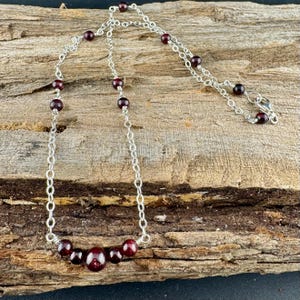 May include: A silver chain necklace with dark red garnet beads. The necklace features a delicate chain design with evenly spaced beads and a larger cluster of beads at the center. The necklace is displayed on a rustic wooden surface.