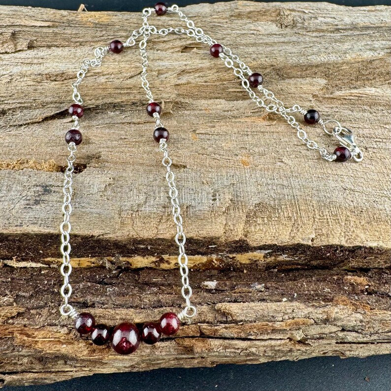 May include: A silver chain necklace with dark red garnet beads. The necklace features a delicate chain design with evenly spaced beads and a larger cluster of beads at the center. The necklace is displayed on a rustic wooden surface.