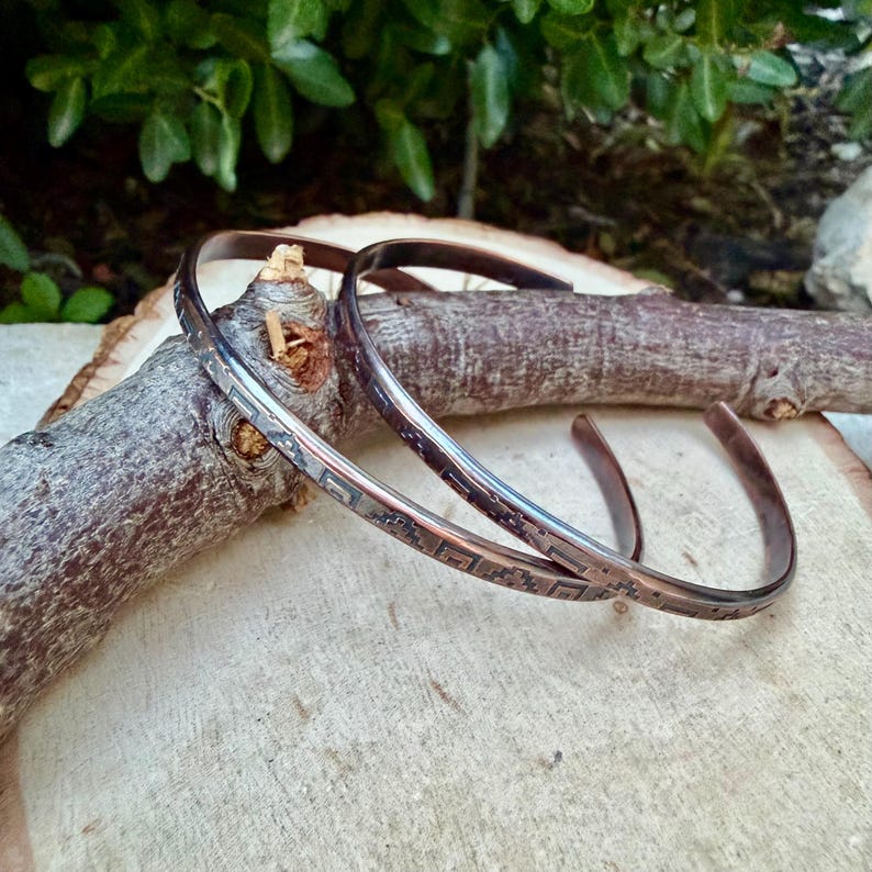 May include: Two thin, open-ended copper bracelets featuring a dark, etched geometric pattern along the outer surface. The metallic bands have a warm, reddish-brown tone and a smooth, polished finish, showcasing a repeating square-based design.