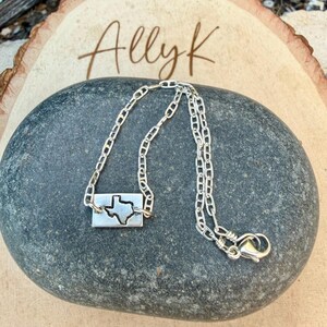 May include: A silver necklace featuring a rectangular pendant with a cutout of the state of Texas. The chain is delicate and has a lobster clasp closure. The word "Allyk" is written in cursive on a wooden surface in the background.
