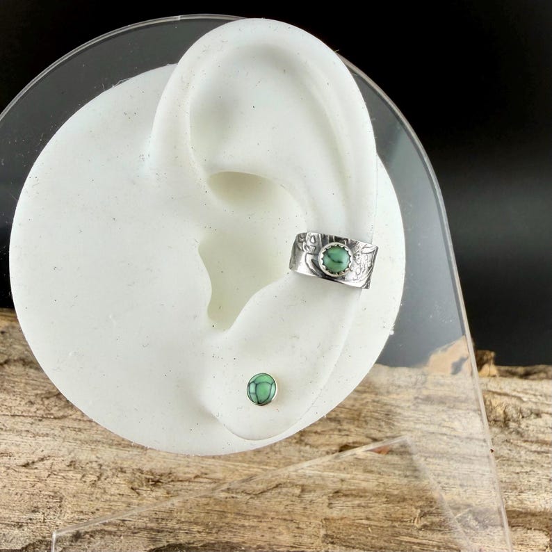 May include: A silver ear cuff and stud earring set. The ear cuff features a turquoise-colored stone set in a decorative band. The stud earring has a matching turquoise stone. The jewelry is displayed on a white ear model.