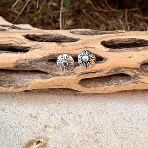 May include: A pair of circular silver stud earrings featuring a raised sun motif with radiating flame-like rays. The metal has a darkened, oxidized finish in the recessed areas to highlight the detailed sun design on each earring.