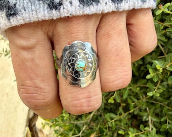 May include: A silver ring featuring a scalloped, circular face with stamped radial patterns and a central, round, light blue and brown matrix turquoise stone. The metal surface displays intricate, repetitive geometric engravings.