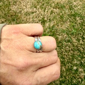 May include: A silver ring with a round, turquoise stone. The ring has a detailed, textured band. The ring is worn on a finger, with a green and brown grassy background.