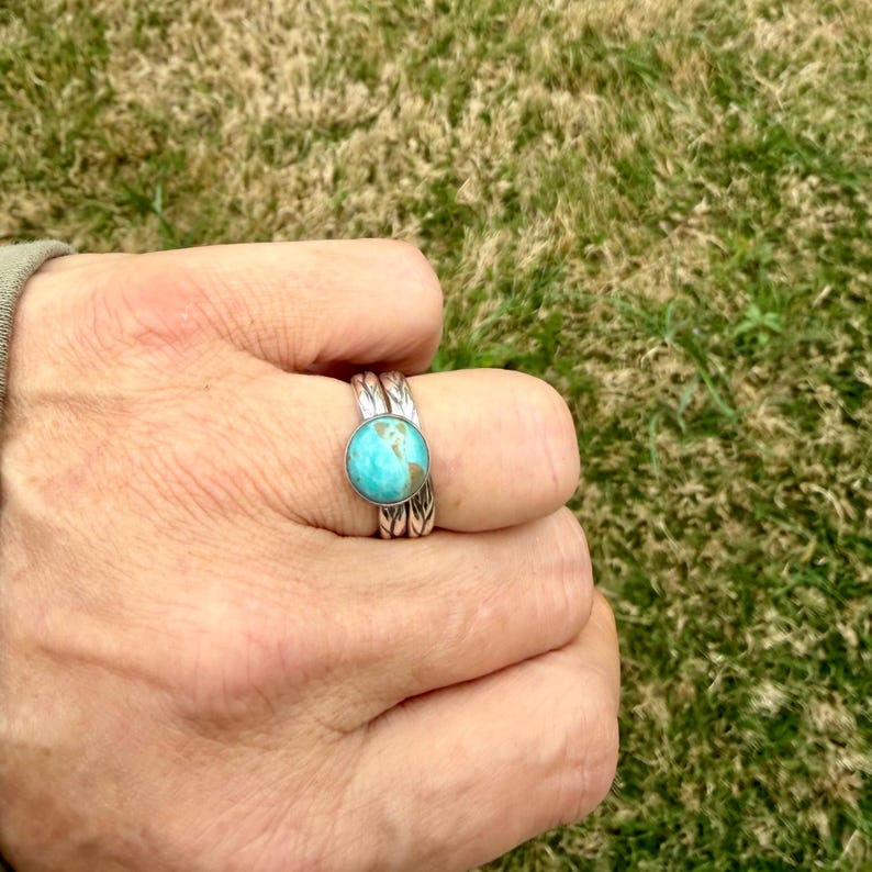 May include: A silver ring with a round, turquoise stone. The ring has a detailed, textured band. The ring is worn on a finger, with a green and brown grassy background.