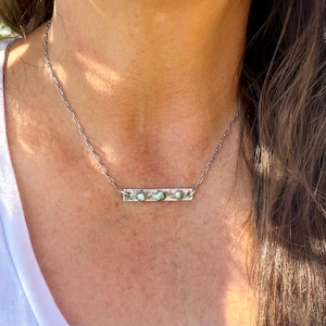 May include: A silver necklace featuring a rectangular pendant with three turquoise-colored stones. The pendant is attached to a delicate chain. The necklace is worn around the neck.
