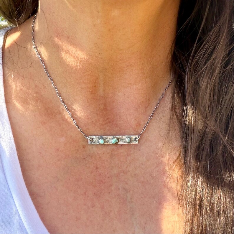 May include: A silver necklace featuring a rectangular pendant with three turquoise-colored stones. The pendant is attached to a delicate chain. The necklace is worn around the neck.