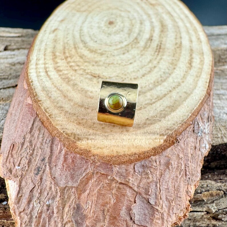May include: A gold-colored ring with a rectangular band and a circular, olive-green gemstone at the center. The ring is displayed on a wooden surface with a natural, textured appearance.
