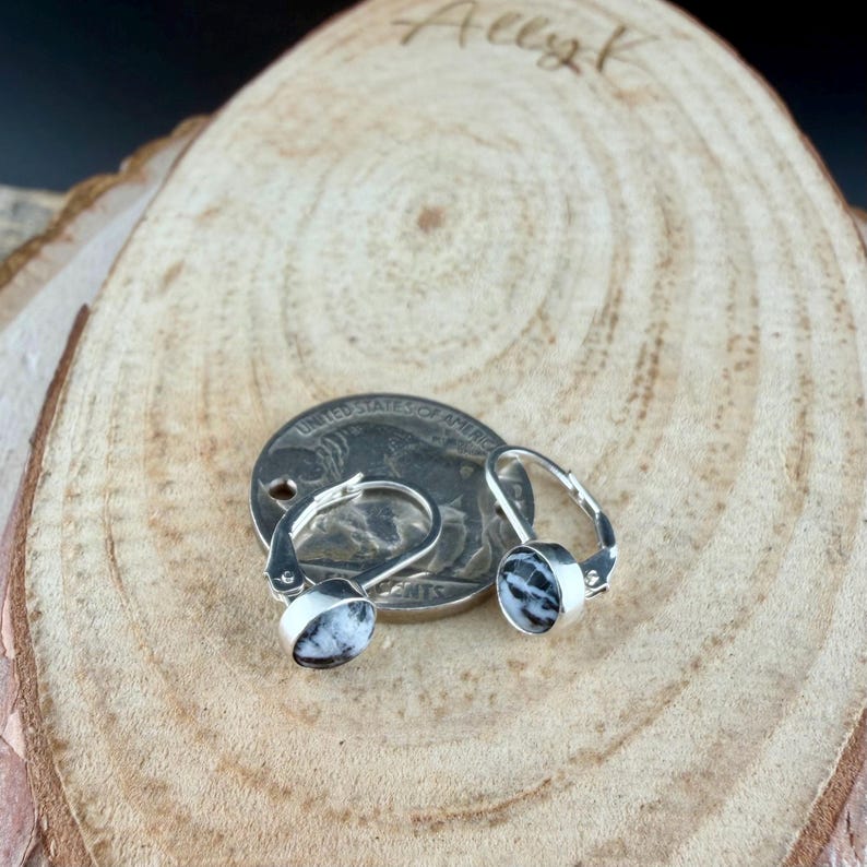 May include: A pair of leverback earrings with round, black and white speckled stones set in silver-colored metal. The earrings are displayed on a wooden surface next to a quarter.