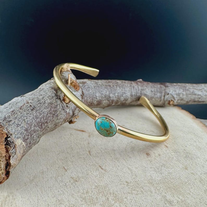 Op de afbeelding: Een goudkleurige manchetarmband met een turkoois steen. De armband heeft een eenvoudig, open ontwerp met een gladde, gepolijste afwerking. De ovale turkoois steen is gezet in een bezel setting, wat een vleugje kleur toevoegt.