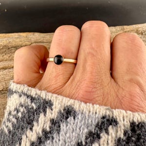 May include: A gold ring featuring a small, round, black gemstone. The ring has a slender band and a minimalist design. The ring is shown on a hand, with a grey and white patterned sweater in the background.
