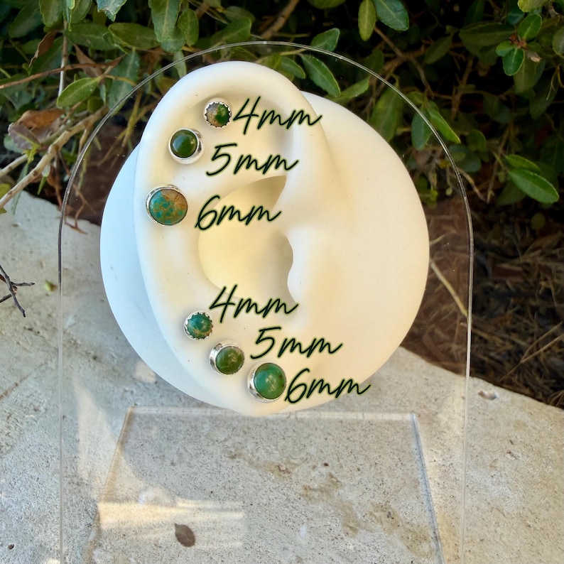 May include: A white ear-shaped display stand featuring six round stud earrings with green and brown stone settings. The earrings are arranged in two rows with text labels indicating sizes of 4mm, 5mm, and 6mm for each corresponding stud.