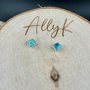May include: A pair of square turquoise stud earrings with silver-colored settings. The earrings are displayed on a wooden surface with the handwritten text "Allyk". The turquoise stones have a light blue color.