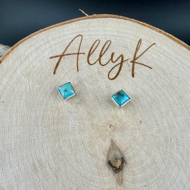 May include: A pair of square turquoise stud earrings with silver-colored settings. The earrings are displayed on a wooden surface with the handwritten text "Allyk". The turquoise stones have a light blue color.