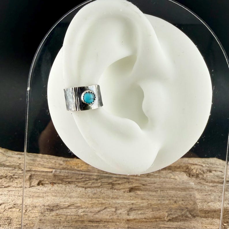 May include: A silver ear cuff with a textured finish and a round, turquoise-colored stone set in the center. The cuff is displayed on a white ear model against a dark background.
