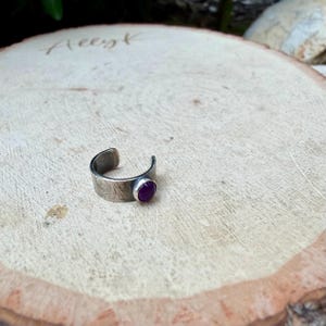 May include: A silver-toned ring with a textured band and a round, deep purple gemstone. The ring is open-ended and sits on a light-colored wooden surface.