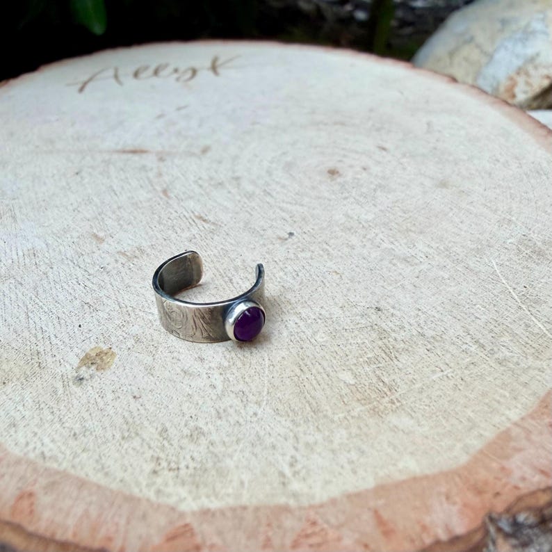 May include: A silver-toned ring with a textured band and a round, deep purple gemstone. The ring is open-ended and sits on a light-colored wooden surface.