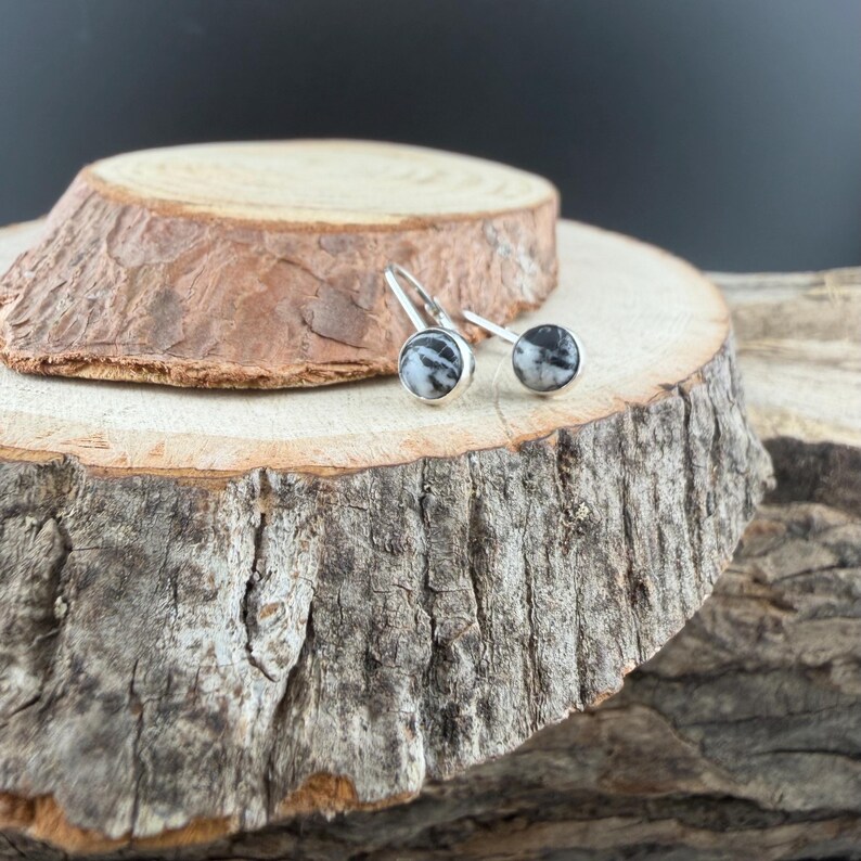 May include: A pair of silver earrings with round, black and white patterned stones. The earrings are displayed on a rustic wooden surface, showcasing their design and texture. The earrings have a simple, elegant design.