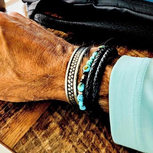 May include: A collection of bracelets on a wrist. The bracelets include a silver woven cuff, a black leather braided bracelet, and a bracelet with turquoise beads. The wrist is against a wooden surface.