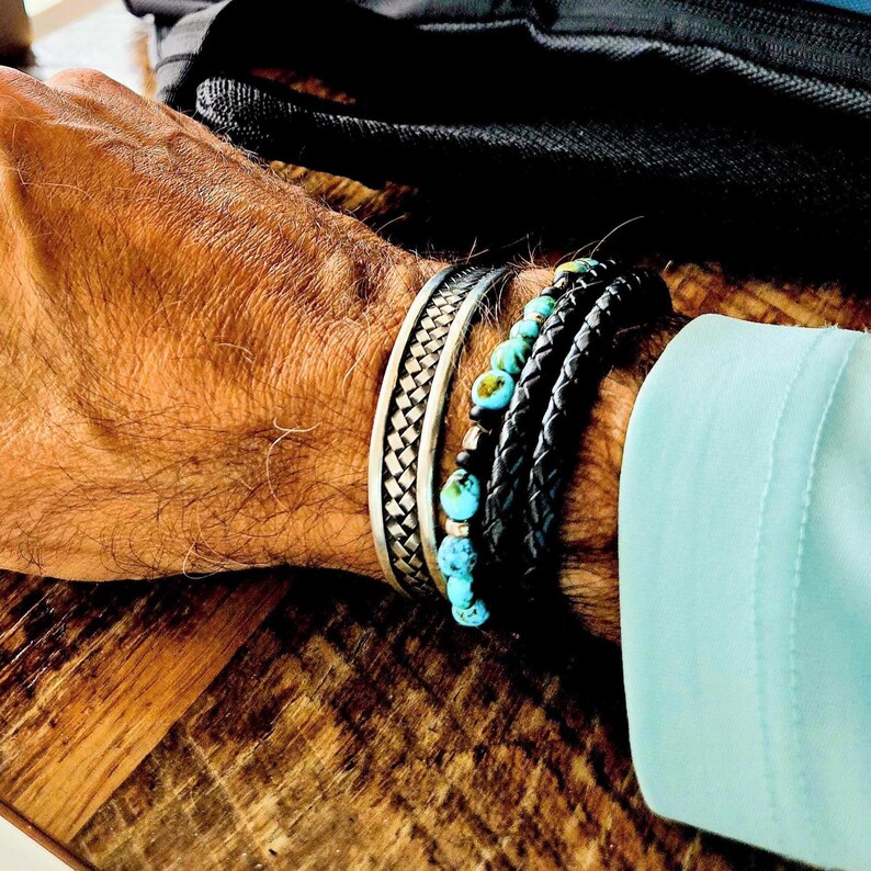 May include: A collection of bracelets on a wrist. The bracelets include a silver woven cuff, a black leather braided bracelet, and a bracelet with turquoise beads. The wrist is against a wooden surface.