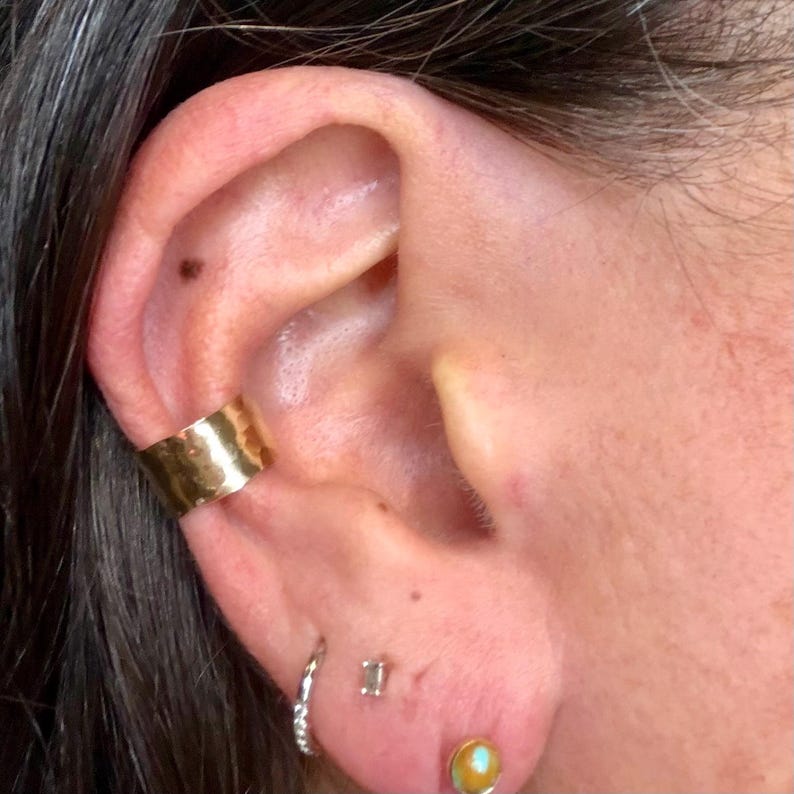 Puede incluir: Primer plano de una oreja adornada con varias joyas. Un brazalete de oreja ancho y dorado est&aacute; en la parte superior de la oreja. Debajo, un peque&ntilde;o pendiente de aro plateado, un peque&ntilde;o perno cuadrado y un pendiente dorado con una piedra redonda y clara son visibles.