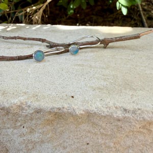 May include: A pair of silver earrings featuring round, iridescent cabochon stones with blue and white hues. The earrings have curved silver ear wires and are resting on a thin, brown wooden twig against a light-colored stone surface.