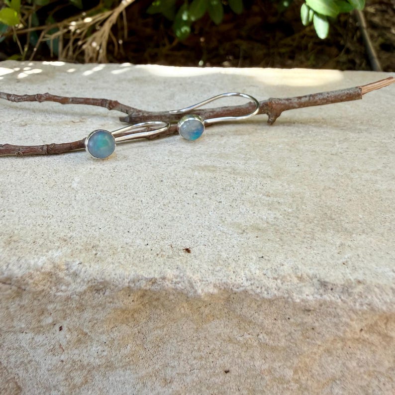 May include: A pair of silver earrings featuring round, iridescent cabochon stones with blue and white hues. The earrings have curved silver ear wires and are resting on a thin, brown wooden twig against a light-colored stone surface.