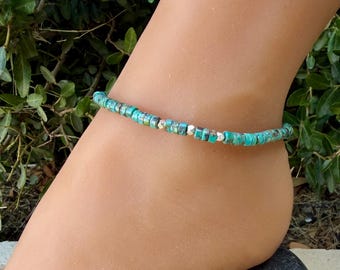 May include: An anklet featuring small, cylindrical turquoise-colored beads with natural dark veining, interspersed with occasional small, metallic silver-toned faceted beads, arranged in a single strand around the ankle.