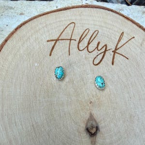 May include: Turquoise stud earrings with a silver-colored border, displayed on a wooden surface. The earrings have a light blue and brown color scheme. The name "Allyk" is written in brown script on the wood.