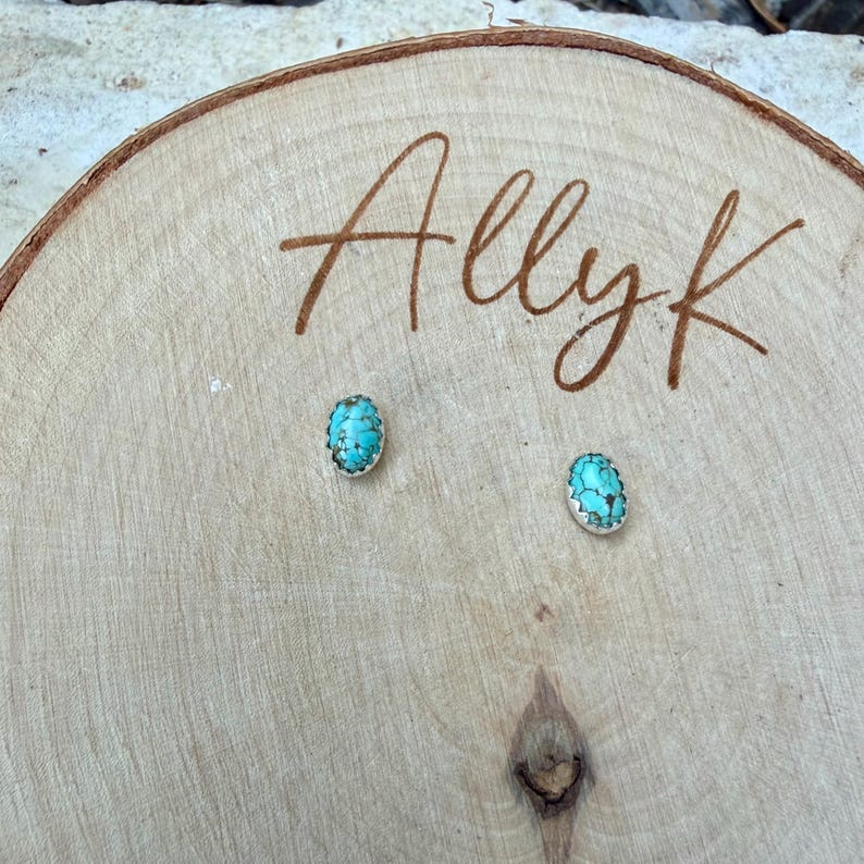 May include: Turquoise stud earrings with a silver-colored border, displayed on a wooden surface. The earrings have a light blue and brown color scheme. The name "Allyk" is written in brown script on the wood.