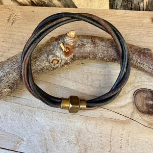 May include: A multi-strand bracelet composed of dark brown and black leather cords twisted together. The bracelet features a central cylindrical brass clasp with a hexagonal shape, providing a secure connection for the leather strands.