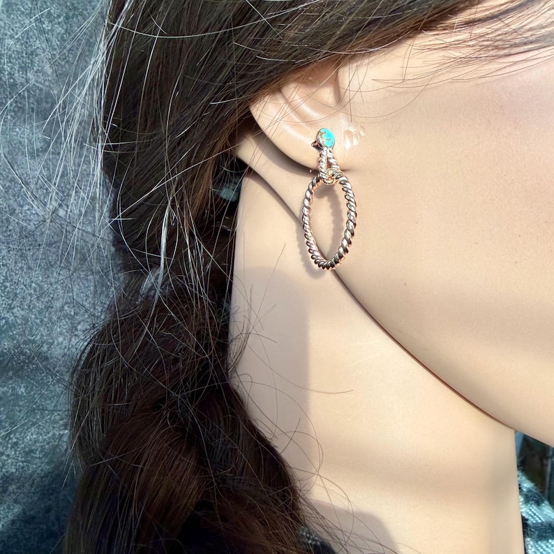 May include: A close-up of a gold-colored earring with a turquoise stone and a twisted rope design. The earring is an oval hoop shape. The earring is worn on a person's ear.
