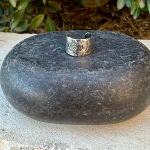 Puede incluir: Un anillo de metal plateado con dise&ntilde;o de banda abierta, textura martillada y un patr&oacute;n geom&eacute;trico de estrella grabado en el centro. El anillo tiene un acabado pulido y muestra un motivo sim&eacute;trico en forma de cruz en su parte frontal.