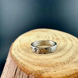 May include: A silver ring with a hammered texture and a cross design. The ring is set on a wooden surface with a dark background. The ring is a simple band style.