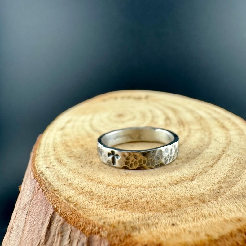 May include: A silver ring with a hammered texture and a cross design. The ring is set on a wooden surface with a dark background. The ring is a simple band style.