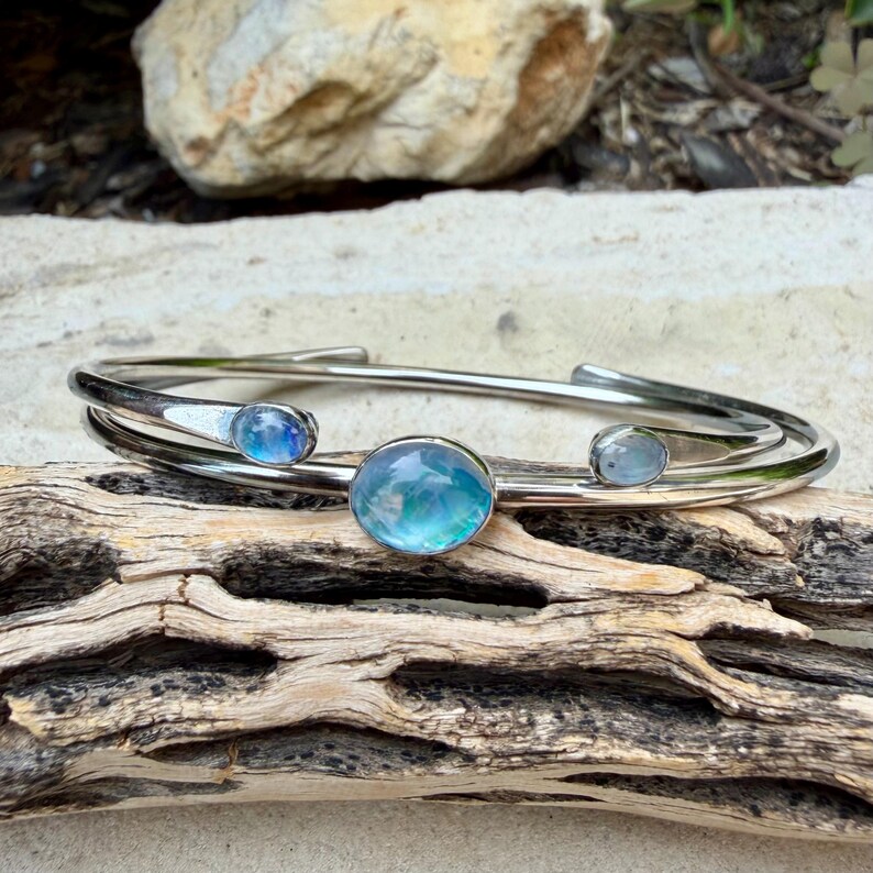 K&ouml;nnte beinhalten: Ein silberfarbenes Armreif-Armband mit drei irisierenden, runden, blau-gr&uuml;nen Edelsteinen. Das Armband hat ein Doppelband-Design und wird auf einem St&uuml;ck Treibholz pr&auml;sentiert. Die Edelsteine haben ein wirbelndes, wasserartiges Aussehen.