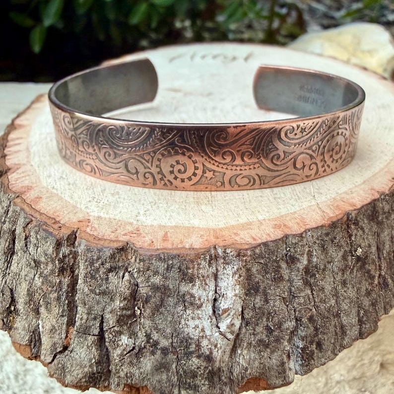 Puede incluir: Brazalete de cobre con un intrincado patr&oacute;n de remolinos. El brazalete abierto tiene un interior oscuro y oxidado y un dise&ntilde;o en relieve detallado en el exterior. El brazalete se exhibe sobre una superficie de madera r&uacute;stica.