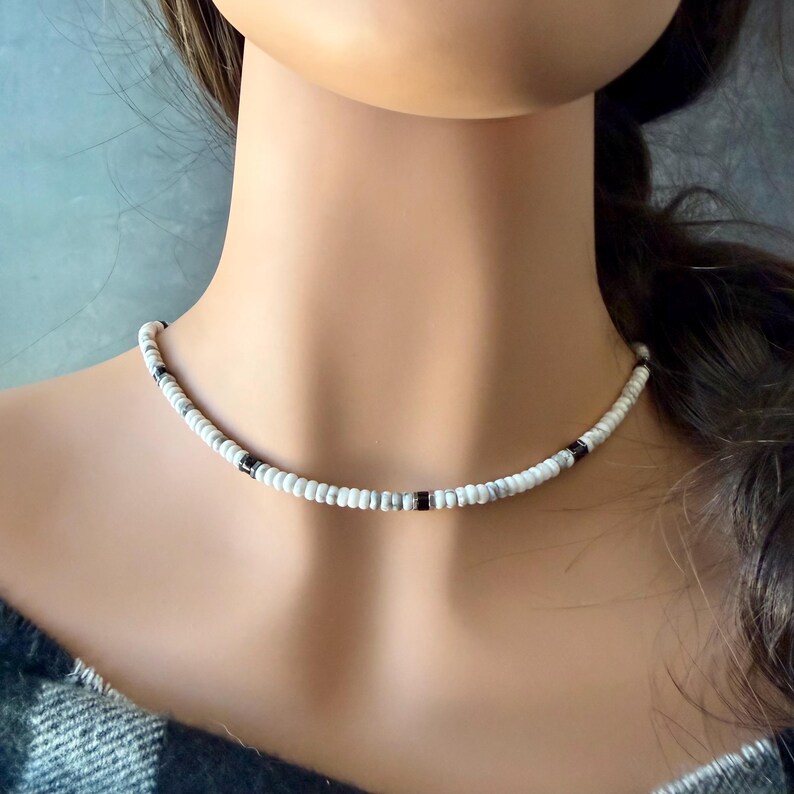 May include: A white beaded choker necklace with black and gray accents. The necklace is composed of small, round beads and is worn around the neck. The beads are strung together to form a delicate, stylish accessory.