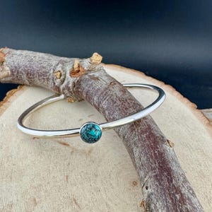 May include: A silver bangle bracelet with a round, turquoise stone set in the center. The bracelet is displayed on a wooden branch against a dark background. The turquoise stone is a vibrant blue-green color.