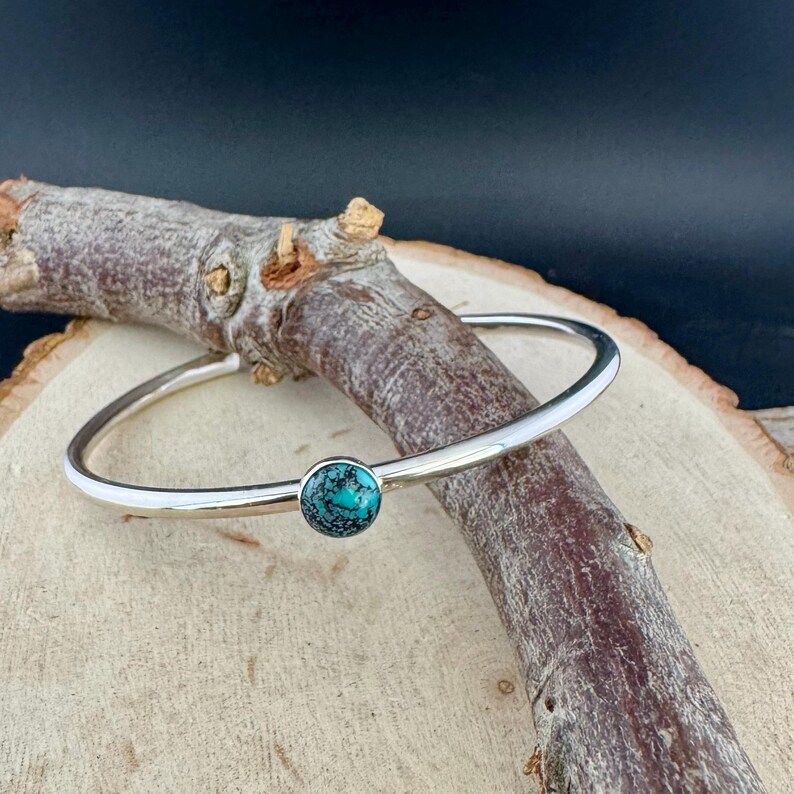May include: A silver bangle bracelet with a round, turquoise stone set in the center. The bracelet is displayed on a wooden branch against a dark background. The turquoise stone is a vibrant blue-green color.