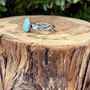 Puede incluir: Un anillo con una piedra de turquesa en forma de l&aacute;grima engastada en un bisel de plata. La banda est&aacute; construida con m&uacute;ltiples hilos de alambre de plata trenzado que se entrelazan para formar una forma circular continua y texturizada.