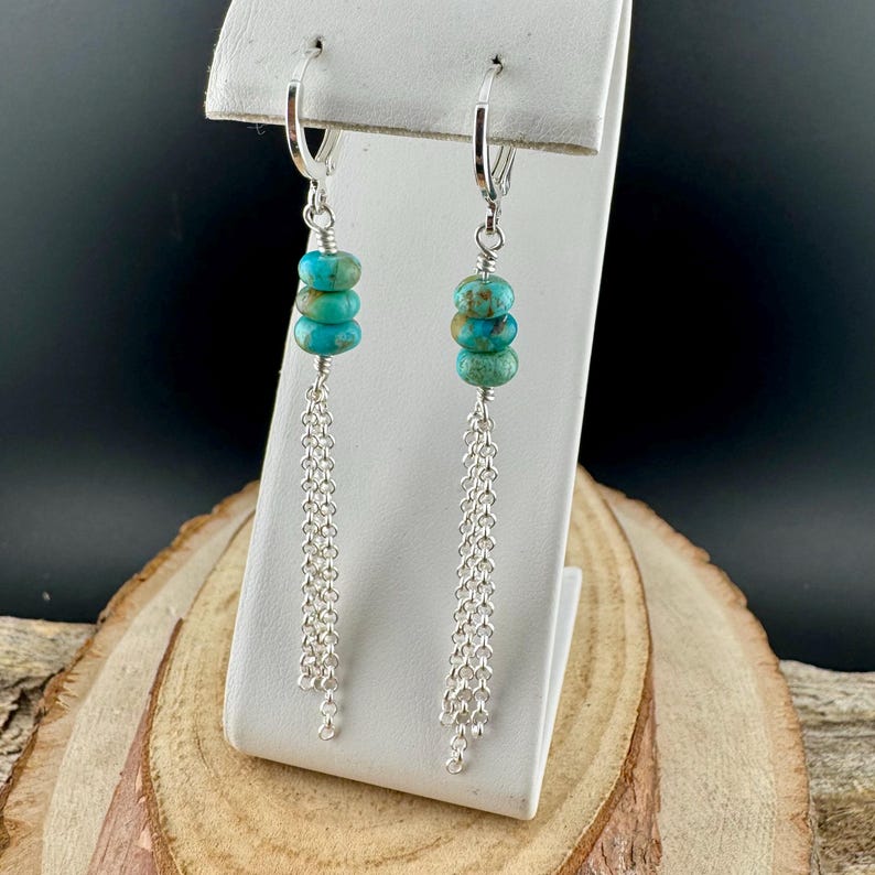 May include: A pair of dangle earrings featuring turquoise-colored beads and delicate silver chains. The earrings have a lever-back closure and are displayed on a white stand against a dark background. The turquoise beads are stacked in a cluster.