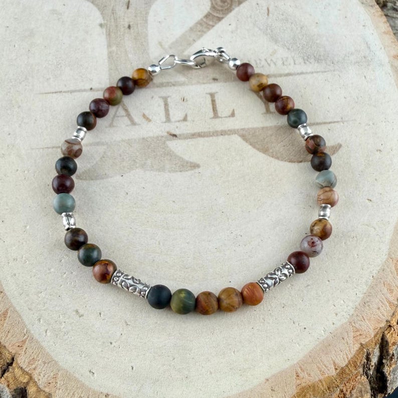 May include: A beaded bracelet featuring round, multi-colored earth-toned stones in shades of brown, green, and grey, interspersed with small silver-toned metal spacer beads and two cylindrical silver-toned metal beads with engraved patterns.