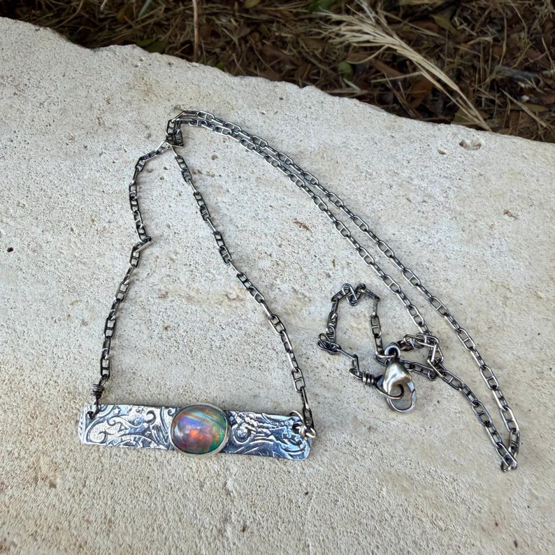 May include: A silver-toned necklace featuring a rectangular bar pendant with an embossed floral pattern and a central oval-shaped iridescent opal cabochon. The pendant is attached to a delicate silver-toned chain with a lobster claw clasp.
