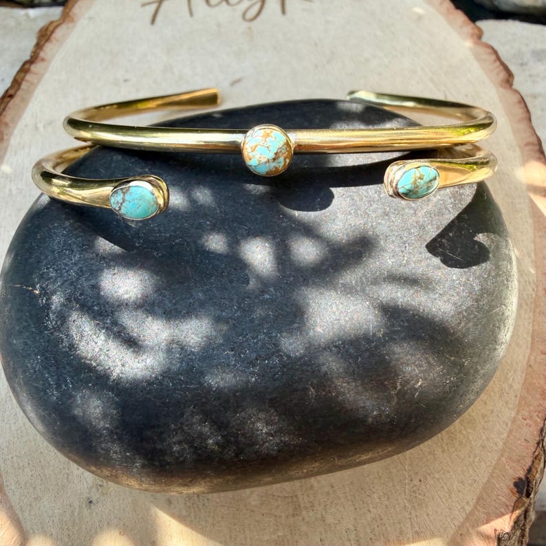 May include: Two gold-colored cuff bracelets with turquoise-colored stones. One bracelet has a single, round stone in the center, while the other has two oval stones on the ends. The bracelets are displayed on a dark gray stone.
