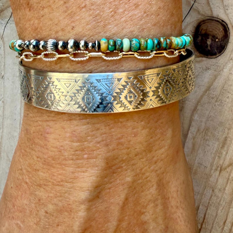 May include: A stack of two bracelets. The top bracelet features a row of small, round turquoise beads and silver-toned metallic beads paired with a gold-toned chain. The bottom bracelet is a wide, silver-toned cuff with an etched geometric pattern.