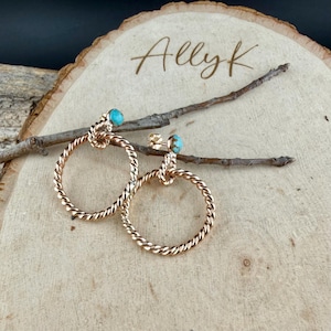 May include: A pair of gold-toned hoop earrings with a twisted rope design. Each earring features a small, round, turquoise-colored stone at the top. The earrings are displayed on a wooden surface with the text "Allyk".
