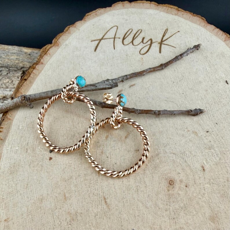 May include: A pair of gold-toned hoop earrings with a twisted rope design. Each earring features a small, round, turquoise-colored stone at the top. The earrings are displayed on a wooden surface with the text "Allyk".