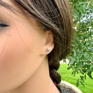 May include: A close-up of a person's ear with a small, round stud earring. The earring has a silver-colored setting and a light blue stone. The background is blurred, with green foliage visible.