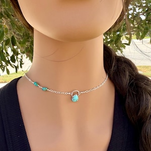 May include: A silver chain necklace featuring a small, teardrop-shaped turquoise pendant suspended from a circular silver link. The chain is accented with three small, round turquoise beads on one side, creating a delicate and balanced design.